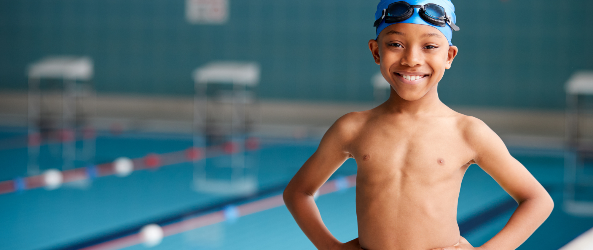 Young boy with blue swimming hat on and green shorts, standing next to an indoor pool